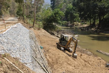 Kırsal inşaat alanında mühendislik projesi için çalışan sarı kazıcı. Nehir kıyısı koruması ve erozyon kontrolü için toprak hareket eden makine kaya seti inşa ediyor.