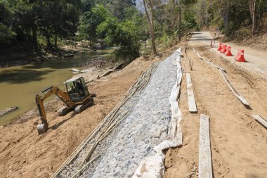 Yol kenarı inşaatı ve mühendisliği, nehir erozyonu kontrolü için jeotextile kumaşlı çakıl döşemesi inşa eden bir kazıcı ile çalışıyor.