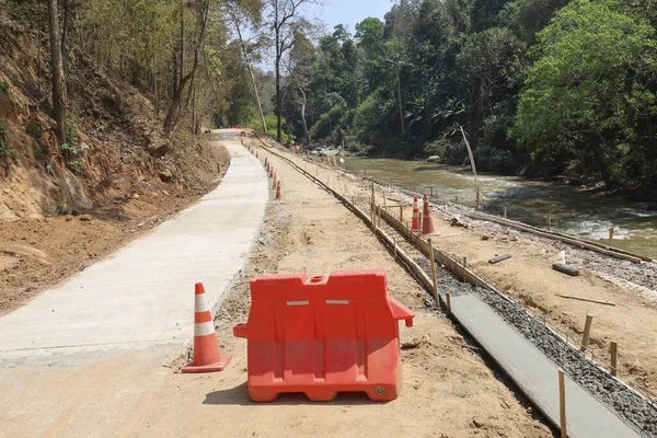 Bereketli yeşil ormanlarda akan nehrin yanında yeni beton yol inşaatı projesi. Parlak gökyüzünün altındaki toprak yolda turuncu bariyer ve güvenlik konisi olan geliştirme alanı
