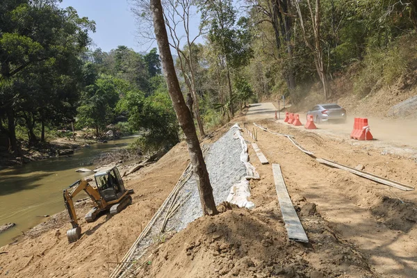 Kırsal yol inşaatı ve nehir kıyısında bir kazıcı. Manzaralı dağ manzarasında eğik dengeleme ve altyapı gelişimi için inşaat mühendisliği çalışmaları