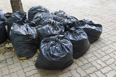 Çöplerle dolu büyük bir yığın siyah plastik çöp torbası kaldırımda toplanmayı bekliyor. Çöp öğütme ve çevre kirliliğinin karmaşık bir sahnesi