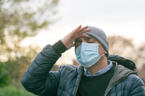 A man in a medical mask in the park with his hand on his head due to a headache. Protection against coronavirus COVID-19 SARS-CoV-2 infection. Selective focus