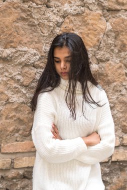 Kollarını kavuşturup tuğladan duvara yaslanan genç, romantik bir esmer kadının portresi. Açık havada.