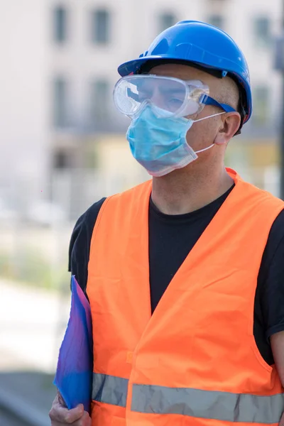Occupational safety and health and protection against adverse conditions at work. Construction worker wearing blue hard hat, reflective vest and protective surgical mask