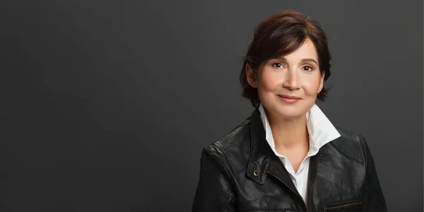 Middle aged well looking woman portrait. Woman wearing white shirt and black leather jacket against dark solid background. Studio female portrait. Billboard size.