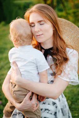 A young mother hugs and soothes her beloved son while sitting on the green lawn. clipart