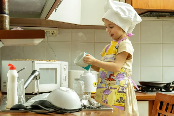 Niña en sombrero de chef blanco está lavando platos en la cocina . — Foto de Stock