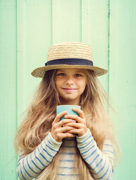 Мила дівчинка стоїть біля бірюзової стіни в капелюсі човна і тримає чашку. Простір для тексту — стокове фото