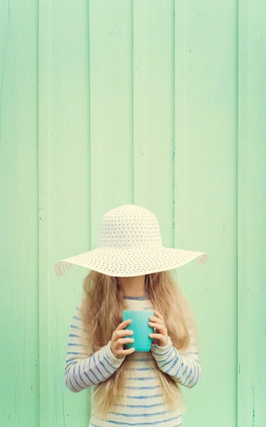 Мила дівчинка стоїть біля бірюзової стіни в білому капелюсі і тримає чашку. Простір для тексту — стокове фото