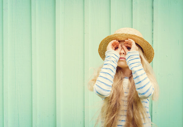 Cute little girl stands near a turquoise wall in boater hat and  looks invented binoculars. Space for text
