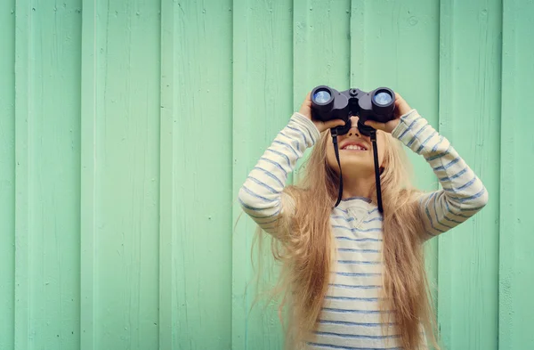 Cute little girl stands near a turquoise wall and looks binoculars. Space for text — Stock Photo, Image