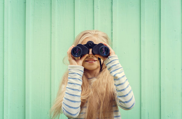 Cute little girl stands near a turquoise wall and looks binoculars. Space for text