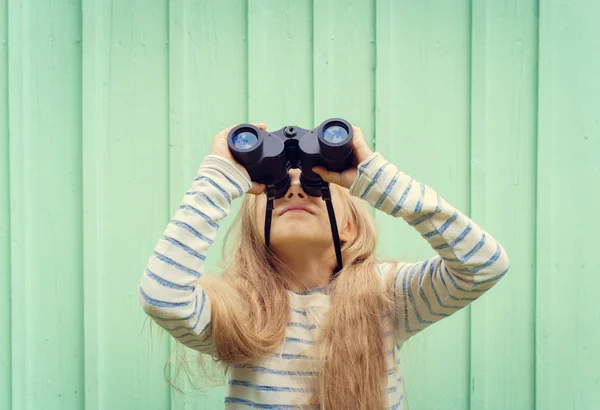 Cute little girl stands near a turquoise wall and looks binoculars. Space for text — Stock Photo, Image