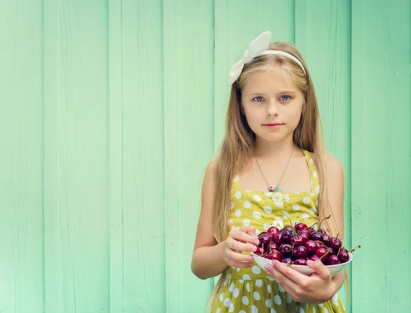 Hermosa chica rubia sobre un fondo de pared de color turquesa placa de retención con cereza . — Foto de Stock