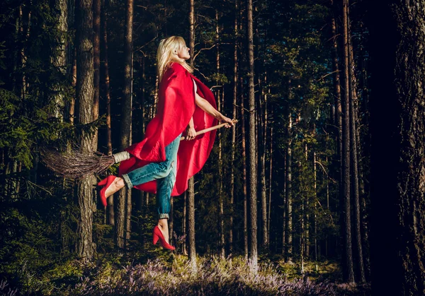 Modern witch blond in jeans and red cape flying on a broom through the forest. Levitation. Halloween holiday. — Stock Photo, Image