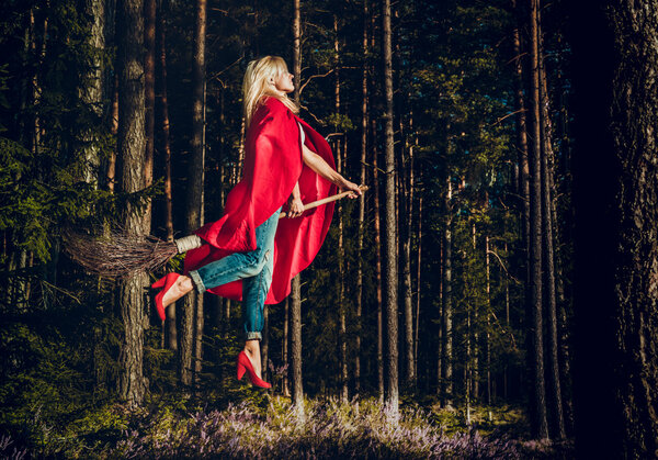 Современная ведьма-блондинка в джинсах и красном плаще летит на метле через лес. Левитация. Хэллоуин
.