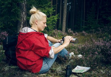 Young active girl tourist drinking tea from a thermos and sitting in the forest. Healthy active lifestyle concept