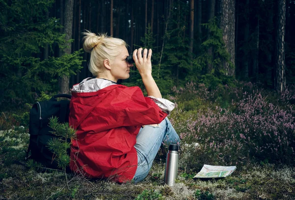 Young active woman tourist sit in a clearing in the woods with a backpack, holding binoculars and a map and looking at the forest. Healthy active lifestyle concept.