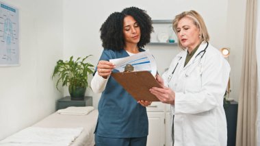 İki doktor klinik muayene odasında hasta bakımı ve tıbbi notları tartışan belgeleri inceliyor. Biri mavi önlük giyerken diğeri beyaz önlüklü, işbirliği ve sağlık ekibinin çalışmalarını ön plana çıkarıyor..