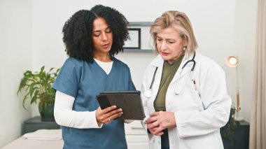 Mavi önlüklü bir hemşire, sakin bir klinikte beyaz önlüklü deneyimli bir doktorla birlikte bir tabletin bilgilerini gözden geçirir..