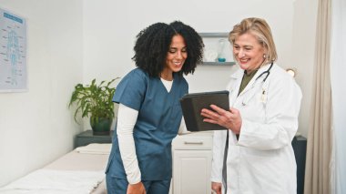 Beyaz önlüklü gülümseyen bir doktor ve mavi önlüklü bir hemşire, modern bir klinikte işbirliği yaparak hasta bilgilerini bir tablet üzerinde gözden geçiriyorlar. Dost canlısı sağlık uzmanları bilgi ve bakımı paylaşıyor.