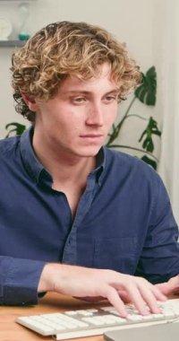 Kıvırcık saçlı mutlu bir genç adam ev ofisinin kurulumunda bilgisayarında özenle çalışıyor, kameraya güvenle gülümsemek için duruyor. Verimliliği ve modern dijital çalışmayı temsil ediyor..