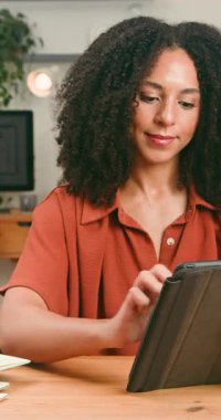 Kıvırcık saçlı mutlu bir kadın çalışmak ya da çalışmak için dijital tablet kullanır. Yukarı bakıyor ve gülümsüyor, üretkenliği ve modern çalışma alanındaki teknolojiyle pozitif bağları aktarıyor..