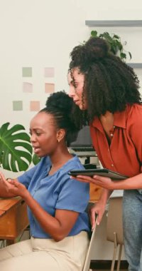 Modern bir ofisteki dijital tabletin içeriğini inceleyen iki farklı iş kadını yakın işbirliği içinde çalışmaktadır. Arkadaşça etkileşimleri ve ortak kahkahaları, takım çalışmasını ve üretkenliği teşvik eden destekleyici ve olumlu bir çalışma ortamını vurguluyor..