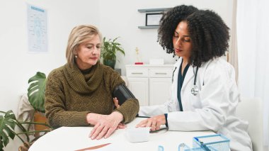 Steteskoplu bir klinisyen, yaşlı bir kadının tansiyonunu temiz bir klinikte ölçüyor. Bakım sahnesinde hasta bakımı, sağlık izleme ve rutin tıbbi ziyaretler vurgulanıyor.