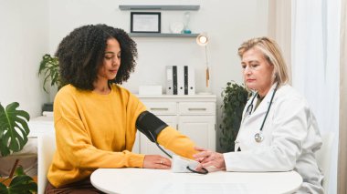 Beyaz önlüklü bir sağlık uzmanı, bir kadının kan basıncını parlak ve temiz bir klinikte kelepçeyle ölçer. Bitkiler ve modern tıbbi ekipmanlarla sakin, şefkatli bir atmosfer..