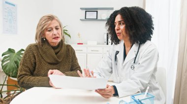 Bir doktor, yaşlı bir hastayla parlak bir klinikte belgeleri inceleyip bakımı tartışıyor. Sıcak, profesyonel ve özenli, hasta-doktor konsültasyon anını yakalıyor..