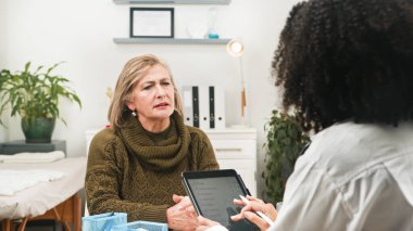 Yaşlı bir kadın, parlak bir klinikte bir klinikle konuşuyor. Tablet ve klasörler masanın üzerinde otururken kahverengi bir süveter giyer ve arka planda raflar ve bitkiler vardır..