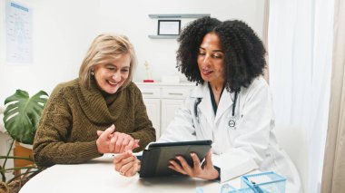 Steteskoplu bir klinisyen, tabletle ilgili bilgileri parlak ve modern bir klinikte gülümseyen yaşlı bir kadına açıklar. Sıcak, profesyonel bakım iletişimi ve hasta ilişkilerini vurguluyor.