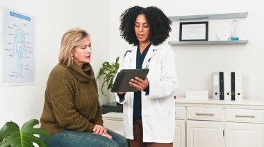 Beyaz önlüklü bir doktor klinikte eski bir hastasıyla konuşuyor, tabletteki bilgileri gözden geçiriyor. Sakin ve profesyonel ortamda tıbbi afişler ve organize raflar yer alıyor..