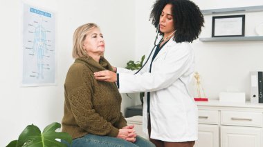 Şefkatli bir kadın doktor, yaşlı bir hastayı, parlak bir klinikte steteskopla kalbini dinleyerek kontrol ediyor. Olay yeri, profesyonel bakım ve tıbbi muayeneyi yansıtıyor..