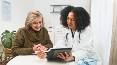 Doktorun, parlak bir klinikteki yaşlı bir hastaya tabletle ilgili bilgileri açıkladığı sıcak bir tıbbi sahne. Profesyonel bakım, güven ve net iletişim.