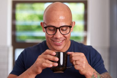 Gözlük takan neşeli, kel bir adam parlak bir mutfakta sıcak bir fincan kahvenin tadını çıkarıyor. Memnuniyetle gülümser, evdeki rahat bir sabahı yakalar..