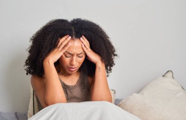 Bir kadın yatakta oturuyor, elleri şakaklarının üzerinde, gözleri acı içinde kapalı. Yumuşak yastıklar ve sakin bir ev düzeni baş ağrısı, stres ve yorgunluk anlarını vurgular.