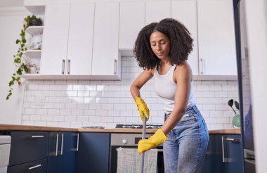 Bir kadın çağdaş bir mutfağı temizliyor, sarı eldivenler ve sıradan kotlar giyiyor, paspas kullanıyor. Parlak beyaz dolaplar, metro fayanslarının geri sıçraması ve doğal ışık yeni ve düzenli bir iç mekan yaratıyor..