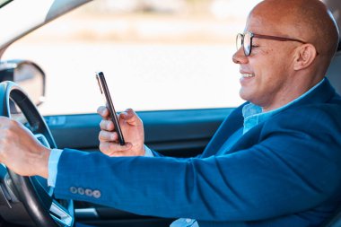 Mavi takım elbiseli neşeli bir işadamı akıllı telefonunu kontrol ederken araba kullanıyor. Görüntü, iş-yaşam dengesi, bağlantı ve iş seyahati sırasında profesyonel güveni taşır.