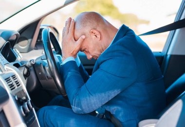 Mavi takım elbiseli profesyonel bir adam arabanın içinde, başı dertteyken oturuyor. Resim yoğun, yorucu bir iş hayatında stresi, tükenmeyi ve anlık bunalımı iletir..