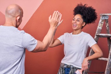 İki arkadaş keyifli bir beşliği paylaşıyor. Bir kadın ahşap merdivenin başında oturmuş, sıcak duvar renkleri ve günlük kıyafetlerle evin tadilatındaki ilerlemeyi kutluyor..