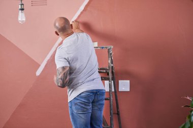 Kotlu ve gri tişörtlü bir adam metal bir merdivenin üzerinde duruyor, pembe bir iç duvar boyuyor. Sahne, yenilenme, odaklanma ve ev yapımı çalışmaları kaydediyor.