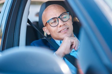 Kendine güvenen, kel bir işadamı gözlük takar ve emniyet kemeriyle arabada oturur. Odaklanmış ve sakin bir şekilde araba sürmeye hazırlanır..