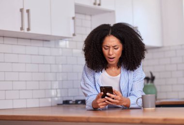 Çizgili gömlekli kıvırcık saçlı bir kadın ahşap mutfak tezgahına yaslanmış, akıllı telefonuna şaşırmış bir ifadeyle odaklanmış. Parlak, modern bir ev mutfağının yanında bir kupa oturuyor..