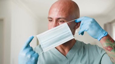 Kendini adamış bir erkek sağlık uzmanı mavi tıbbi eldiven giyerken cerrahi bir maske takıyor. İşe hazırlanıyor, tıbbi bir ortamda güvenlik ve hasta bakımına olan bağlılığını gösteriyor..