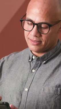 Gözlüklü neşeli kel bir adam akıllı telefonundan kafasını kaldırıp kameraya doğru gülümsüyor. Mutlu ve meşgul görünüyor. Bağlantı ve modern iletişimi gösteriyor..