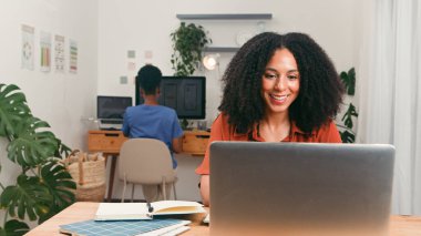 Gülümseyen bir kadın, arka plandaki bilgisayara odaklanırken, modern, temiz bir masada dizüstü bilgisayarda çalışır. Parlak bitkiler, organize alan ve işbirlikçi atmosfer üretkenliği ve yaratıcılığı iletir.