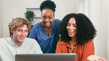 Üç genç profesyonel, parlak, modern bir ofiste, takım çalışması, yaratıcılık ve pozitif, çeşitli bir iş ortamı sergileyen bir dizüstü bilgisayarın başında işbirliği yapıyor..