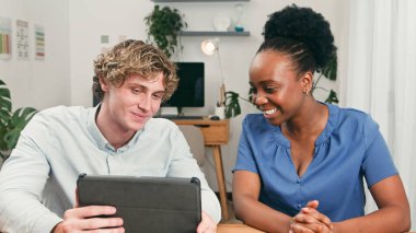 İki farklı takım arkadaşı bir tabletteki içeriği incelerken aynı gülümsemeyi paylaşıyor. Parlak, bitki dolu bir ofis, dostça bir başlangıç atmosferi ve verimli bir işbirliği öneriyor.